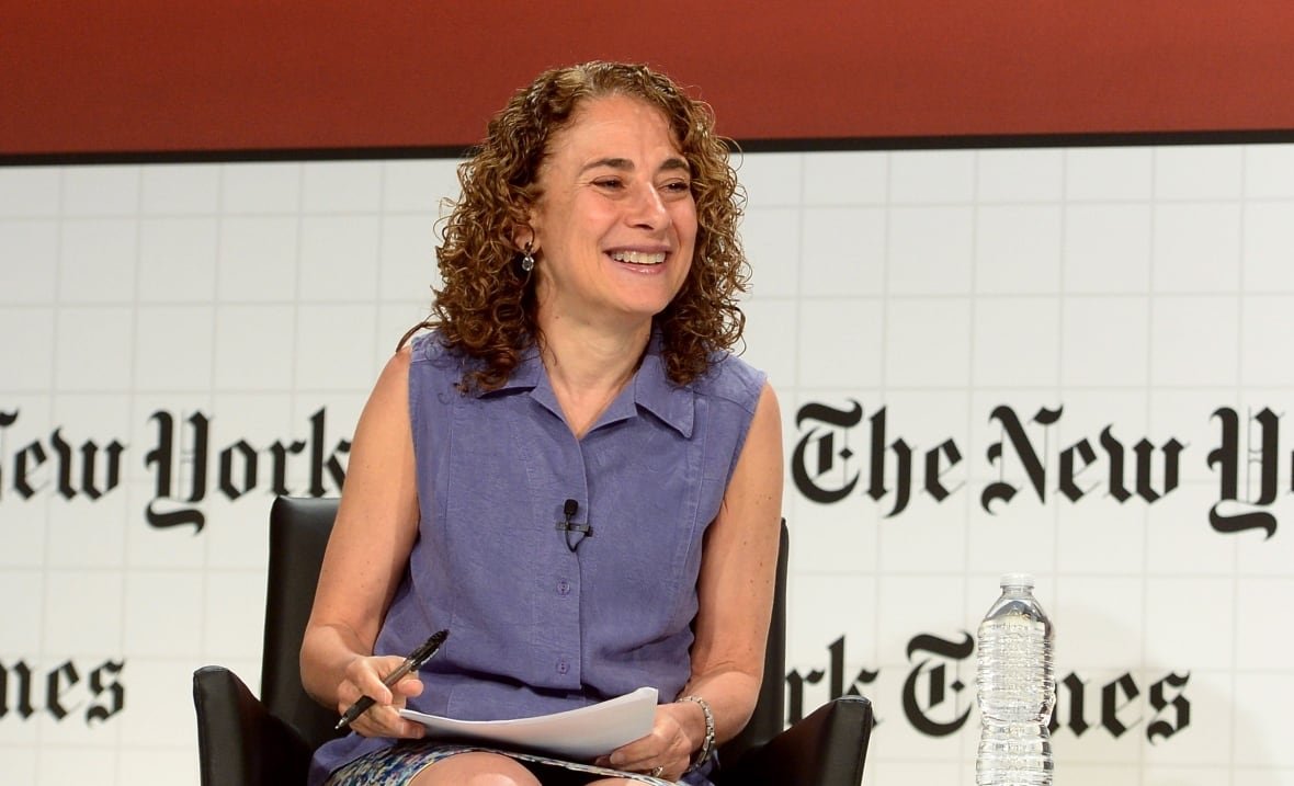 A smiling woman with curly blonde hair sits in a chair on a stage. 