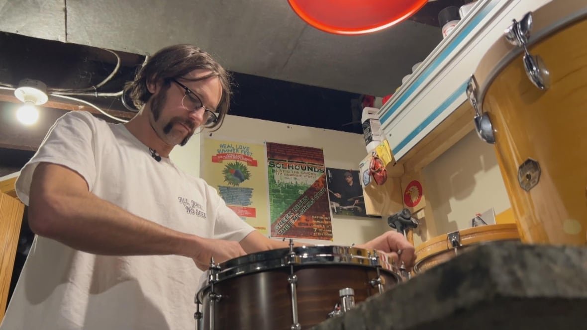 A man stands in front of a drum in a room with posters on the wall behind him.