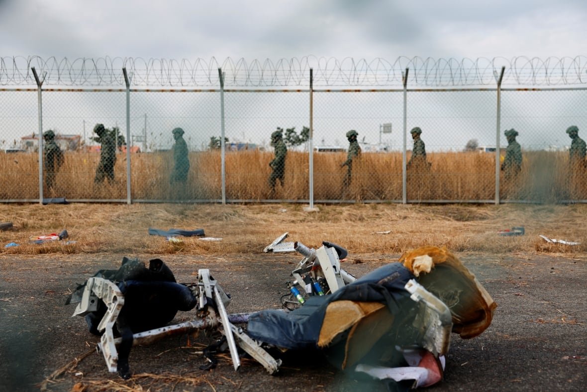 Military personnel walk beside a chain-link fence. In the foreground, debris from an airplane crash is strewn on a runway. 