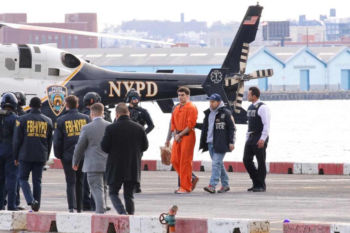 Several police officers escort a man in an orange prison jumpsuit from a helicopter pad. The black-and-white helicopter in the background is marked with the letters "NYPD".