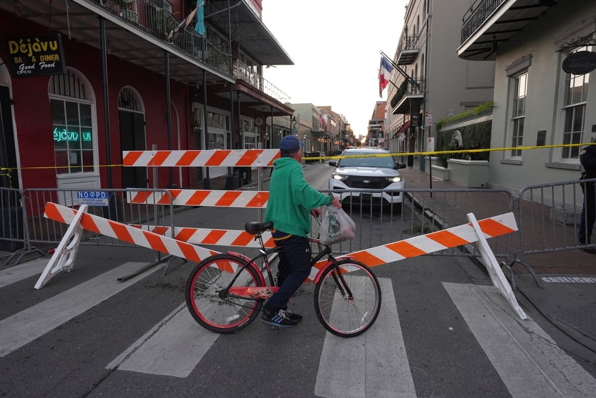 A man on a bike pauses in the street in front of a barricade to look over it down the blocked off street. The barricade is made of metal posts and white and orange striped wooden barriers. Police tape stretches across the street past the barricade. 