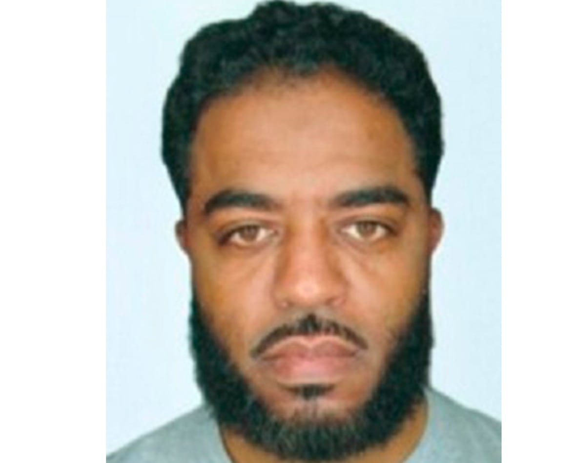 A man with dark hair, a bear and moustache poses for a passport photo. 