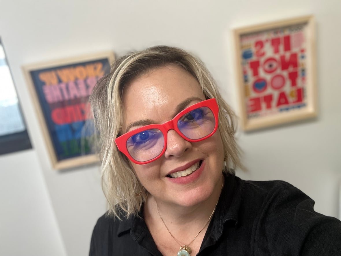 A woman with a blonde bob and red-rimmed glasses wearing a black sweater is smiling at the camera. 