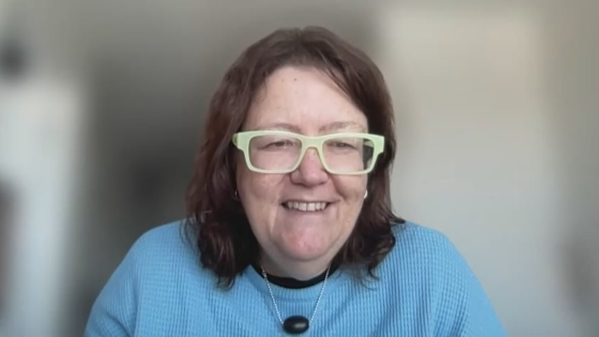 A woman wearing a blue sweater and glasses smiles during a Zoom call.