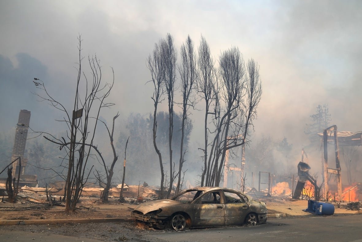 A burned out car is shown parked in front of a burned-down house and several trees that are burned of all vegetation. Orange flames are seen in the background