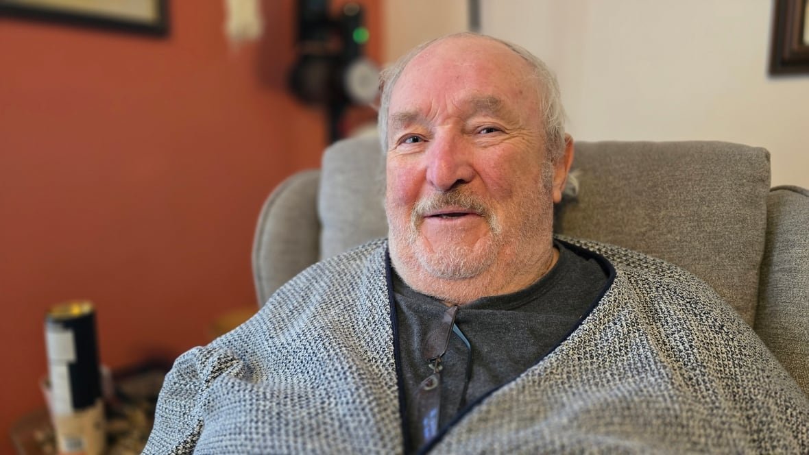 A man sitting in a chair with white hair and a grey and white mustache looks into the camera while wearing a grey sweater.