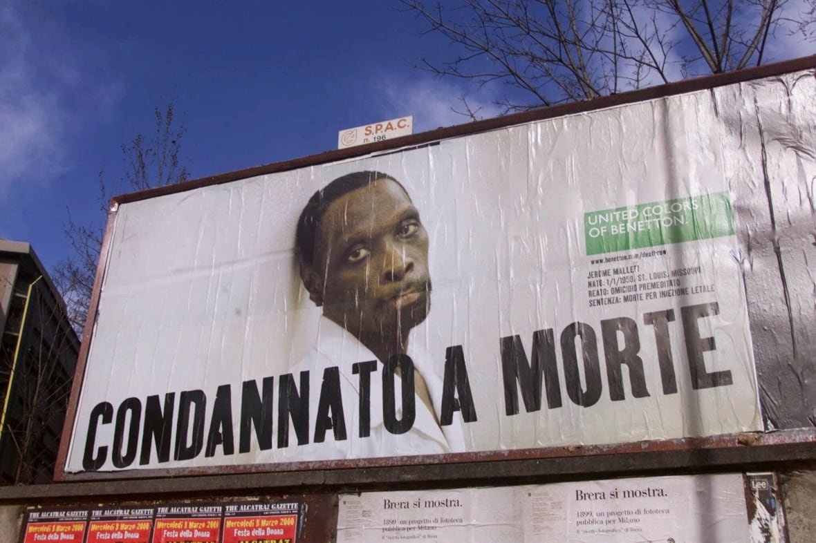 A billboard is shown in closeup. On it, an image of a cleanshaven, dark complected man is shown. He appears forlon. In Italian it reads Condannato A Morte, which translates to Condemned to Death.