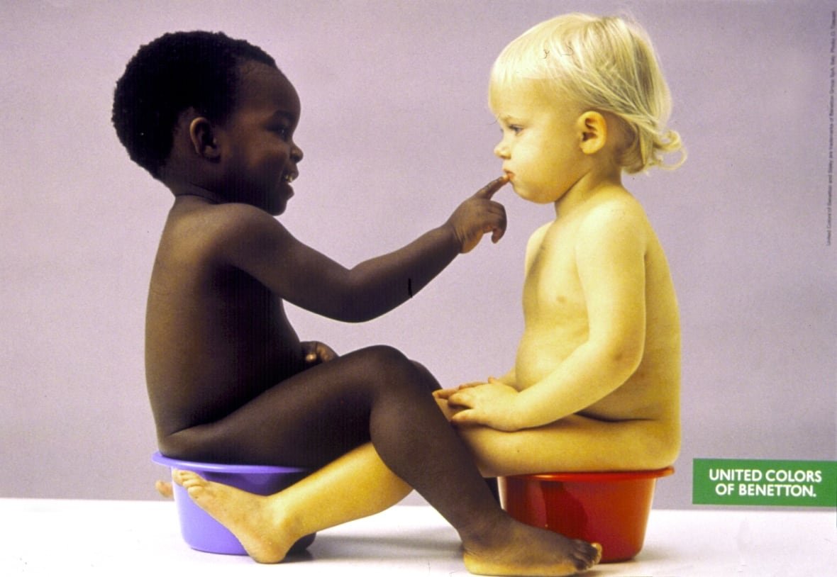 Two young naked boys are shown sitting on portable toilets. A dark complected baby touches the lips of a Caucasian-appearing baby.