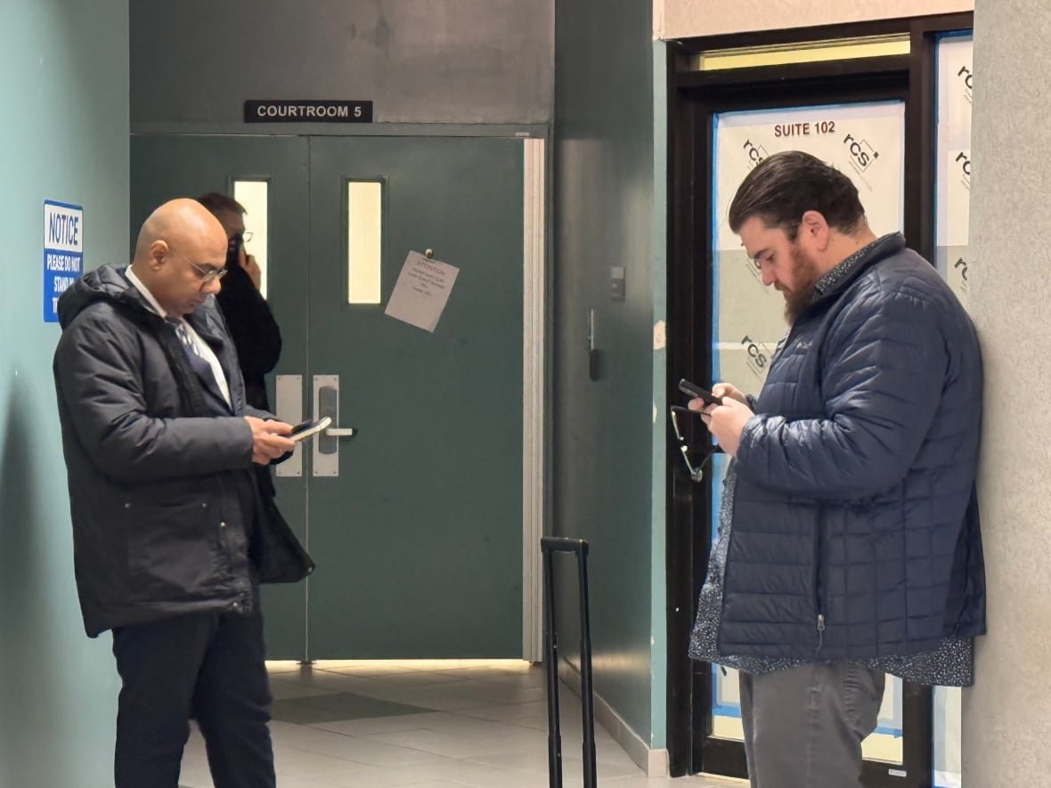 A man wearing a blue coat looks at his phone while holding glasses outside of a courtroom.