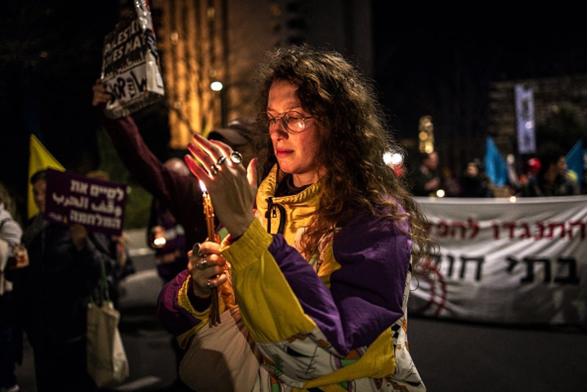 A woman in a yellow and purple jacket holds a candle, using one hand to shield it. Behind her are people holding banners and signs.