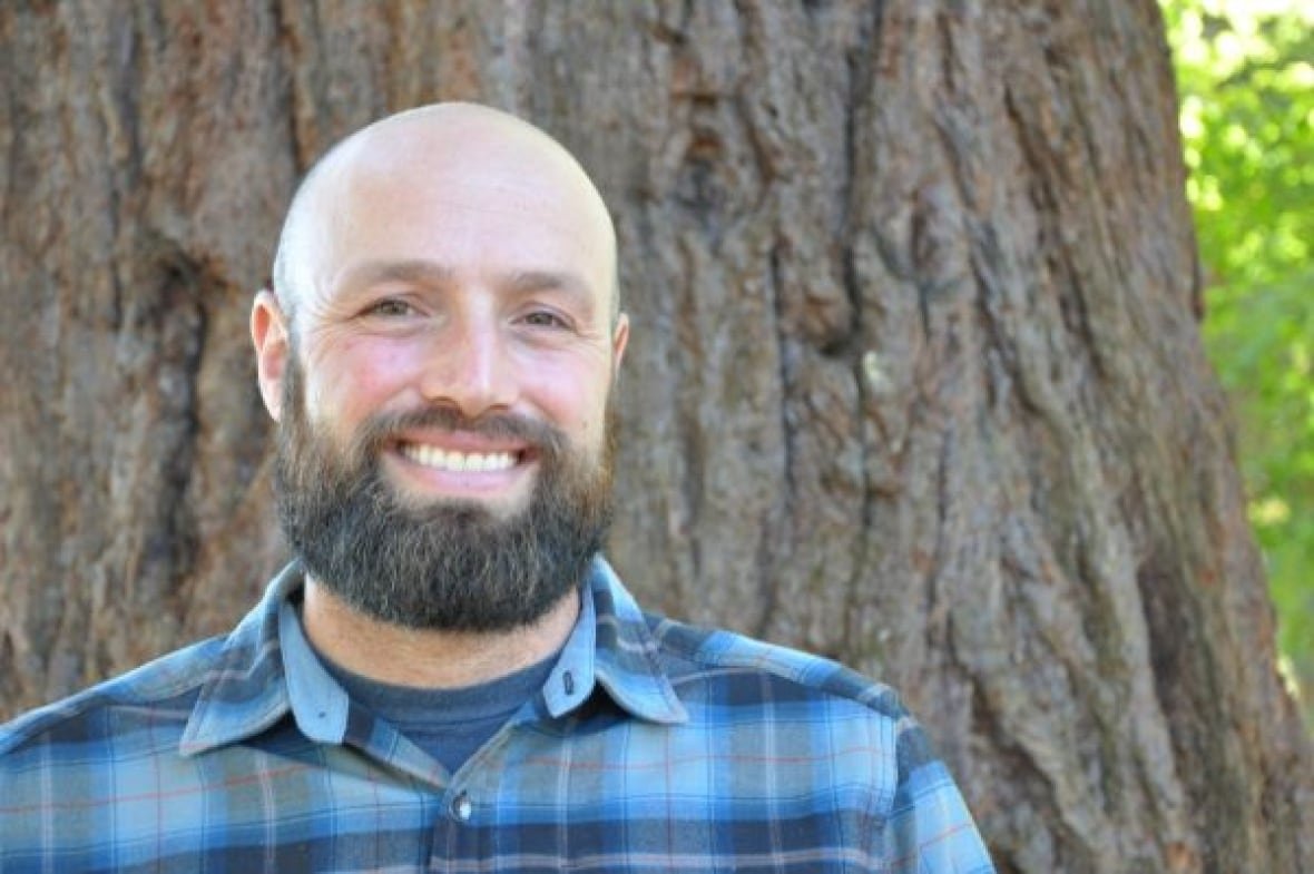 A man with a beard is standing in front of a tree and smiling at the camera. 