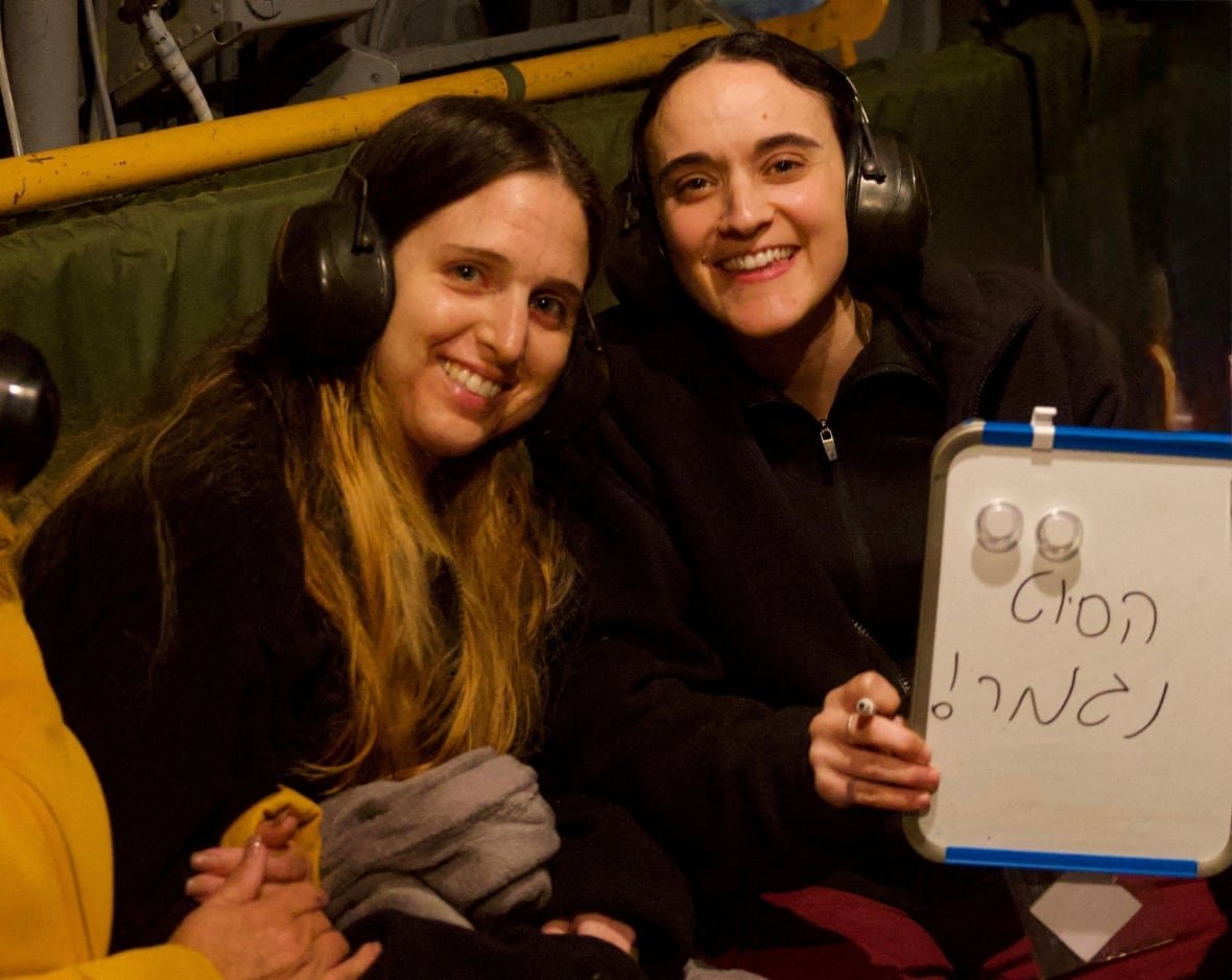 Two women wearing protective ear muffs smile for the camera. One holds up a whiteboard, on which something is written in Hebrew.