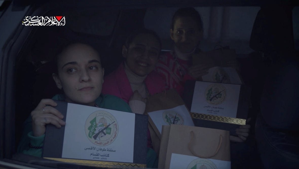 Three women seated together hold up paperwork bearing the logo of Hamas.