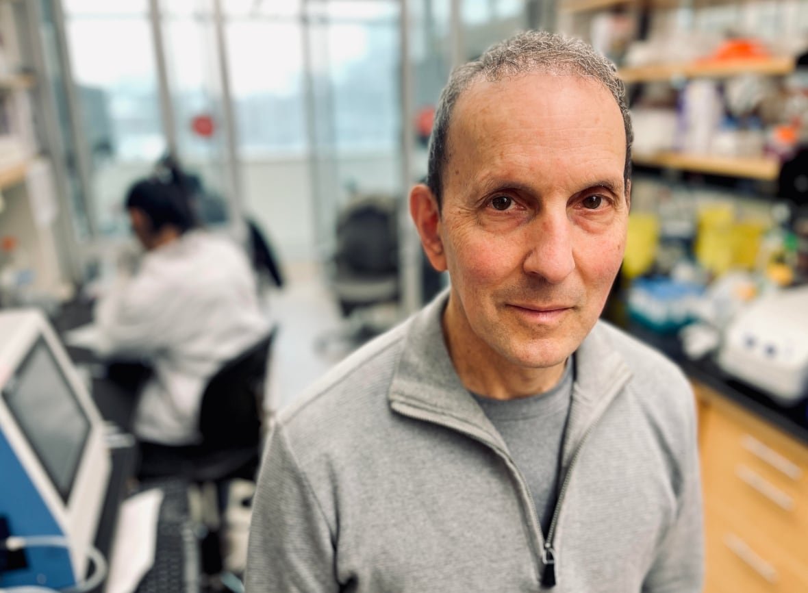 A man with grey hair standing in a lab wearing a grey sweater with lab workers in background out of focus. 