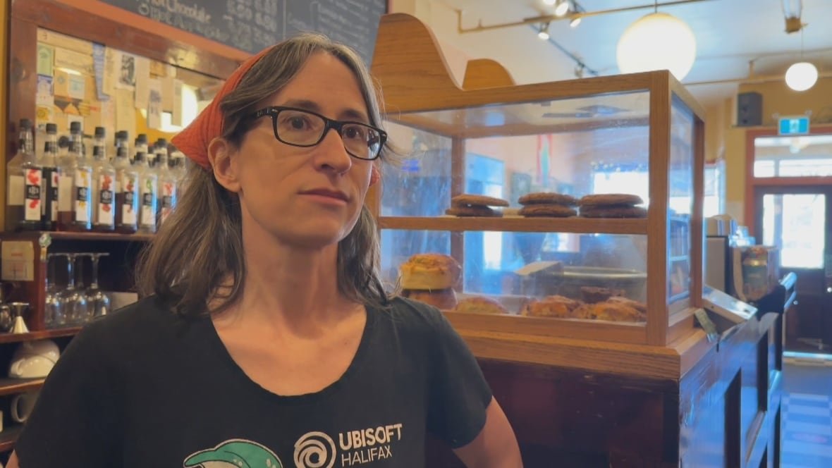 A white woman is wearing glasses and has her brown hair tied back with a red bandana. She is standing in a cafe.