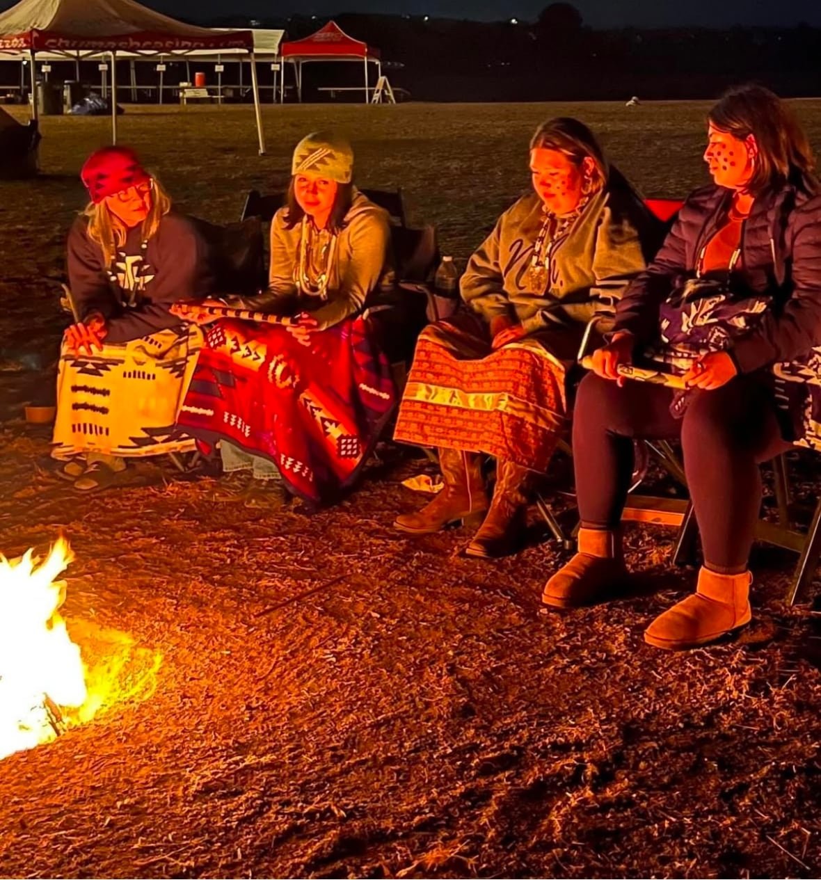 Four women gather around a fire wearing skirts and wrapped in blankets to keep warm.