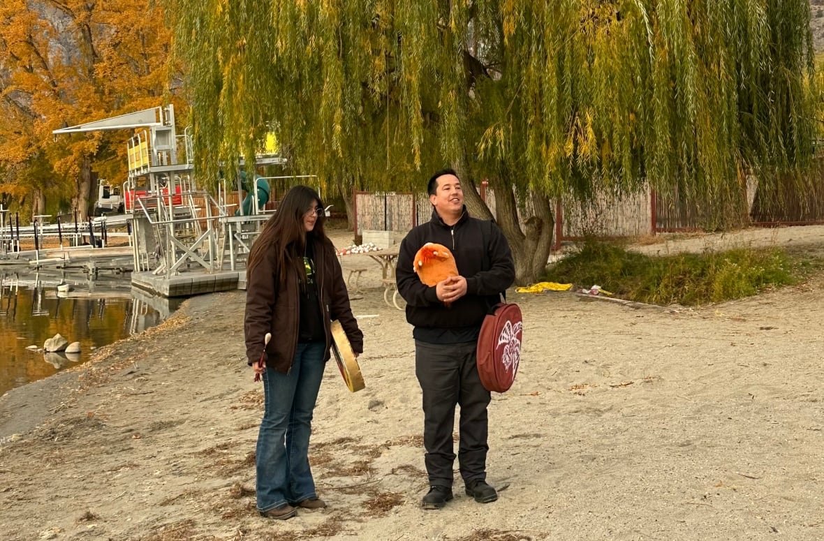 Man and daughter stand near the shoreline, holding hand drums about to sing a welcoming song. 