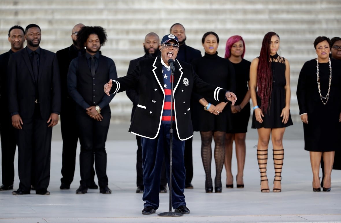 A singer gestures while performing outdoors as a line of people stand behind.