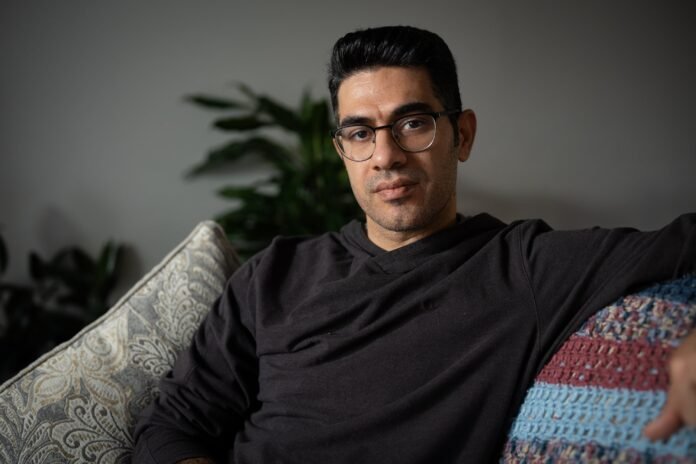 A man in a black shirt and glasses sits on a couch.